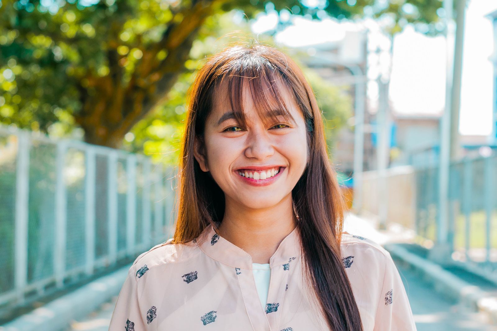 Positive Asian female with long hair looking at camera with smile while standing on sunny street with trees and metal fences on blurred background
