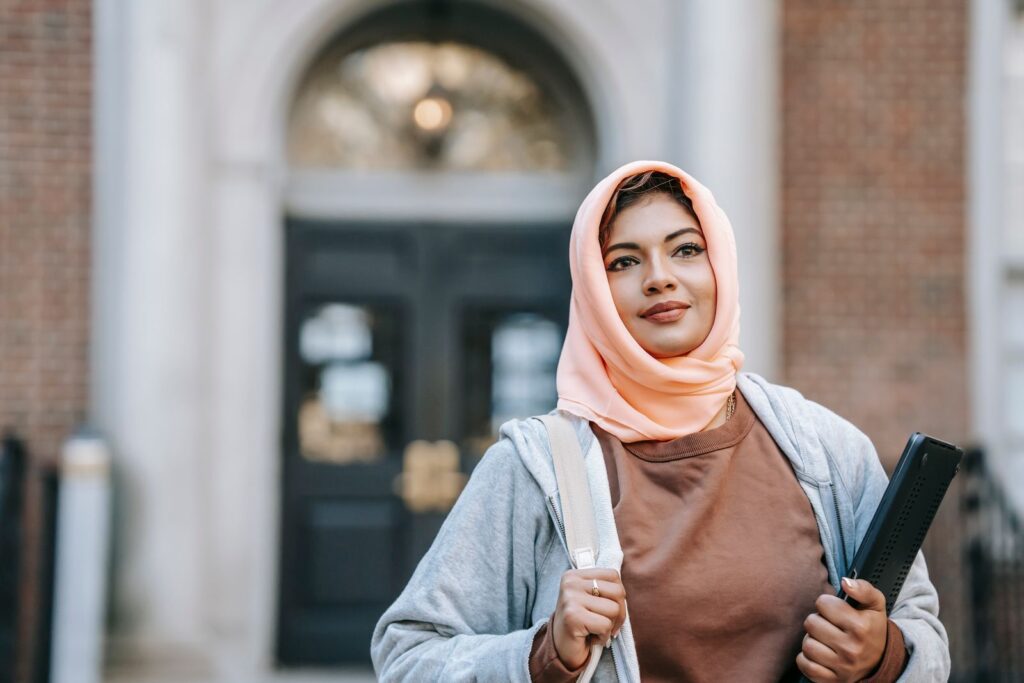 Positive Muslim woman in casual outfit and traditional headdress with laptop on street