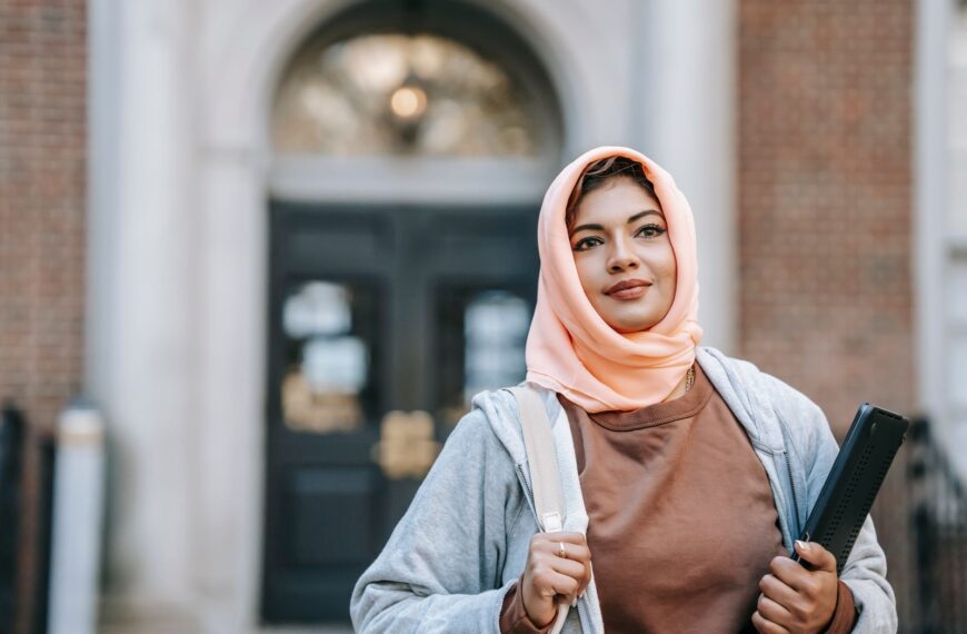 Positive Muslim woman in casual outfit and traditional headdress with laptop on street