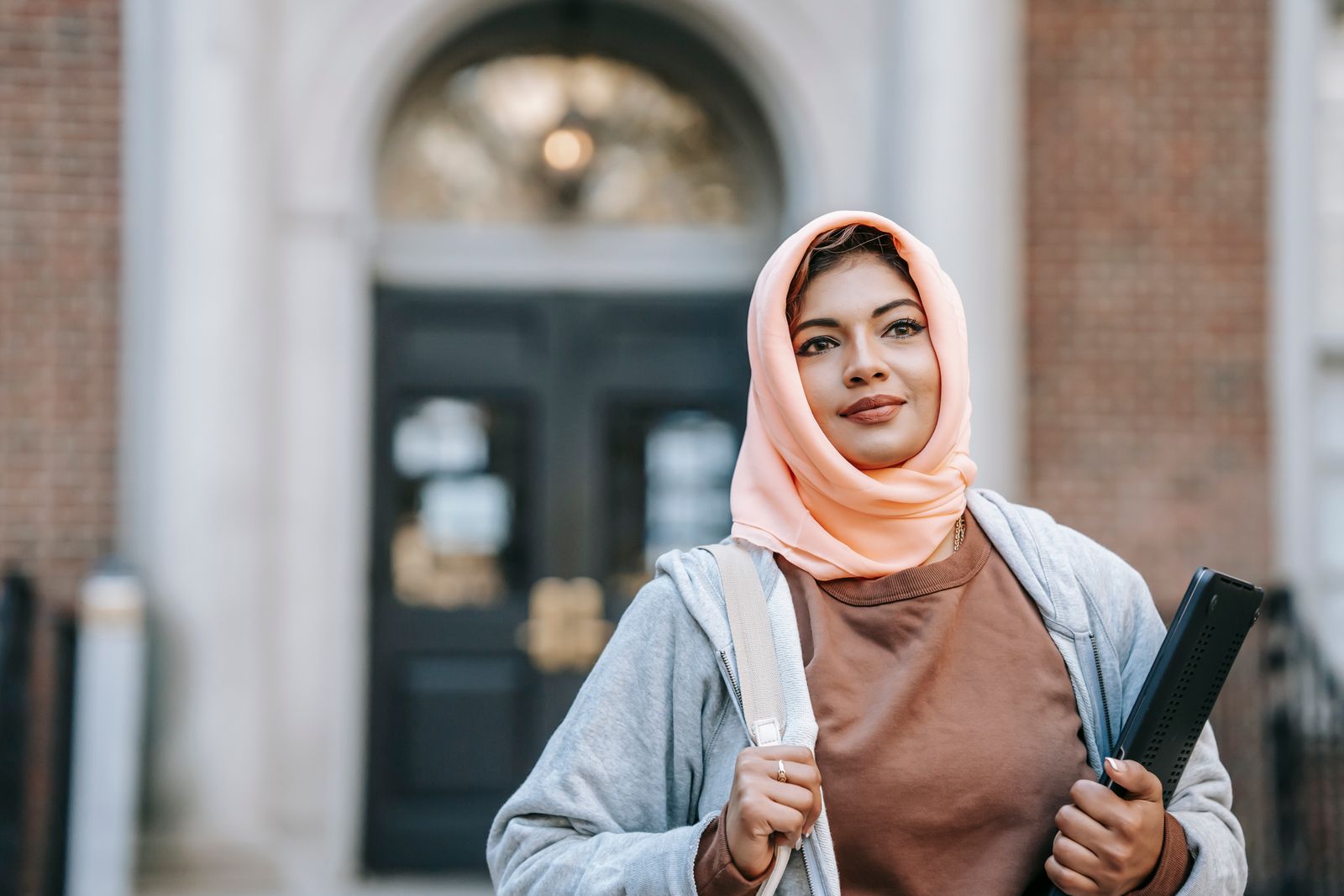 Positive Muslim woman in casual outfit and traditional headdress with laptop on street