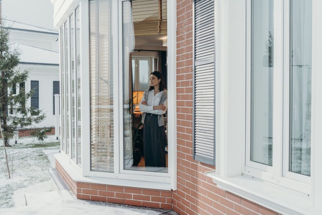 A woman stands indoors, arms crossed, looking out large windows on a snowy day, reflecting indoors and outdoors contrast.