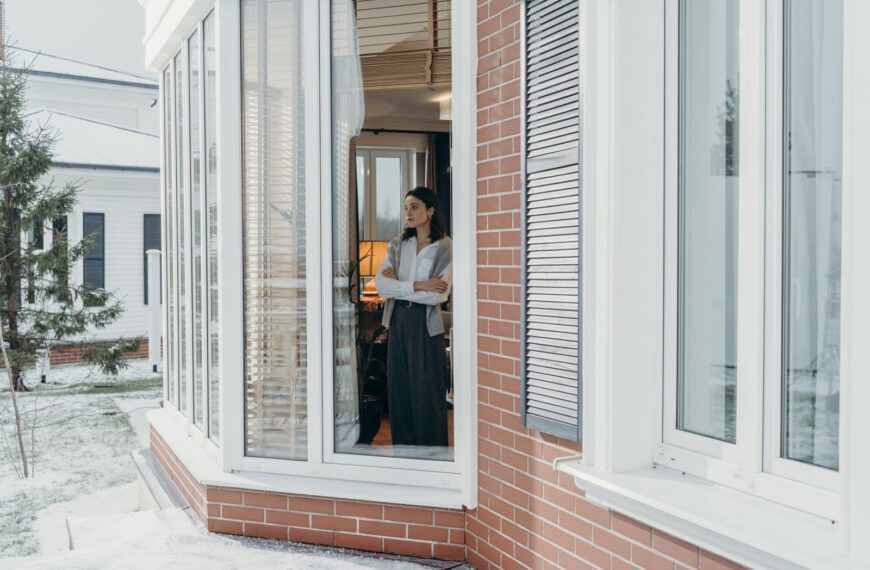A woman stands indoors, arms crossed, looking out large windows on a snowy day, reflecting indoors and outdoors contrast.
