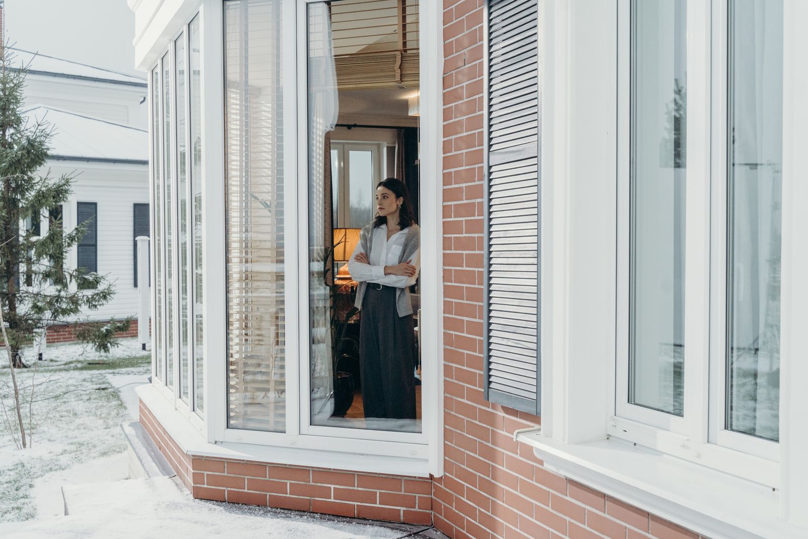 A woman stands indoors, arms crossed, looking out large windows on a snowy day, reflecting indoors and outdoors contrast.