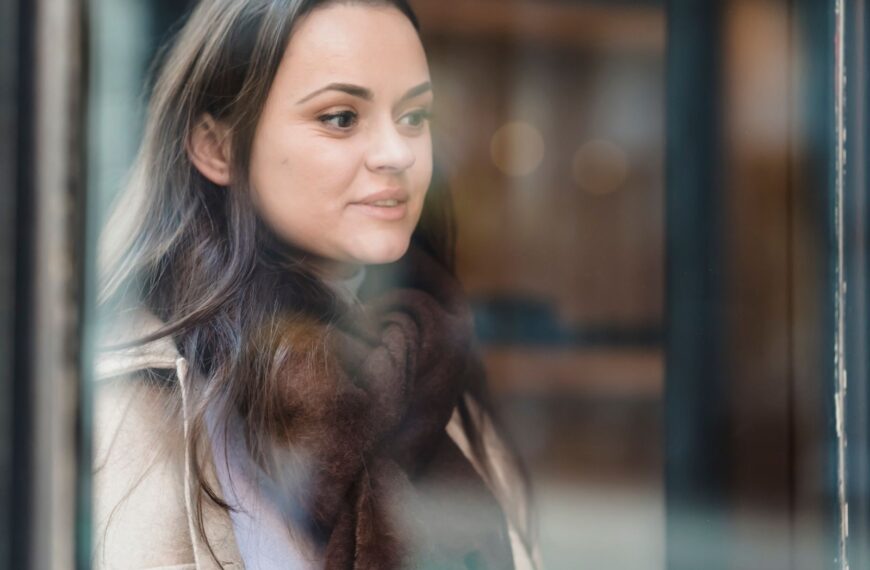 Through glass of crop positive female wearing warm outerwear standing near glass wall and looking away
