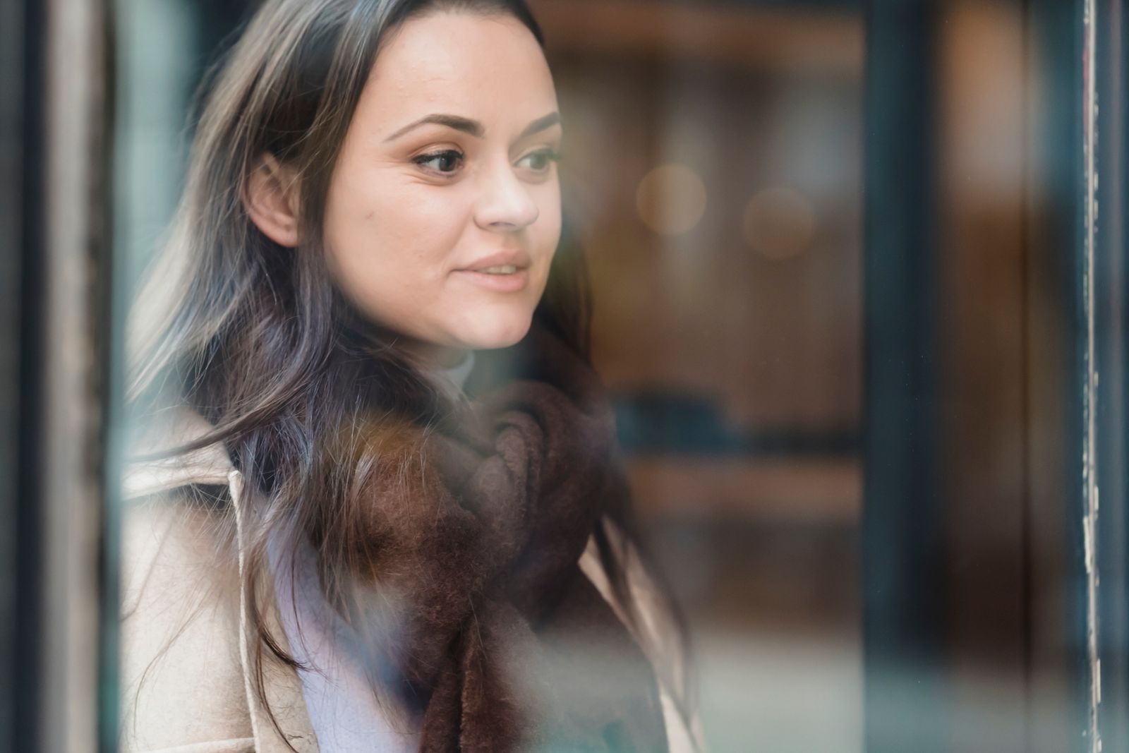 Through glass of crop positive female wearing warm outerwear standing near glass wall and looking away