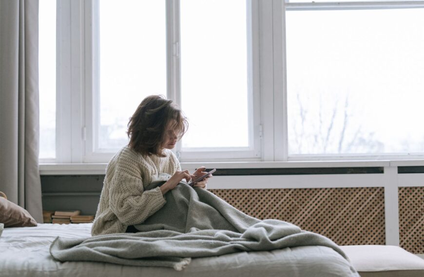 A woman in a knitted sweater using her smartphone while sitting on a bed with a blanket.