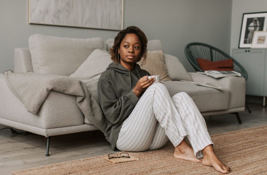 Woman in a gray hoodie drinking coffee while sitting on the floor in a cozy living room.