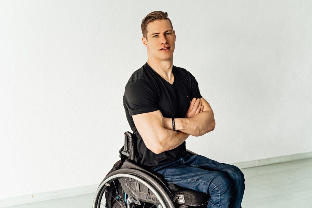 Portrait of a confident young man sitting in a wheelchair indoors with crossed arms.