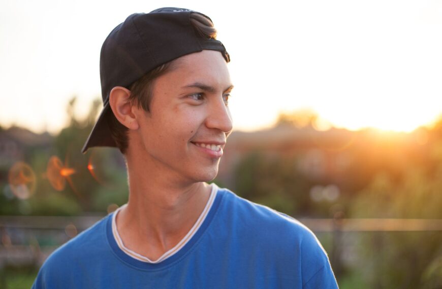 Portrait of a smiling young man in a blue shirt and cap, enjoying a sunset outdoors.