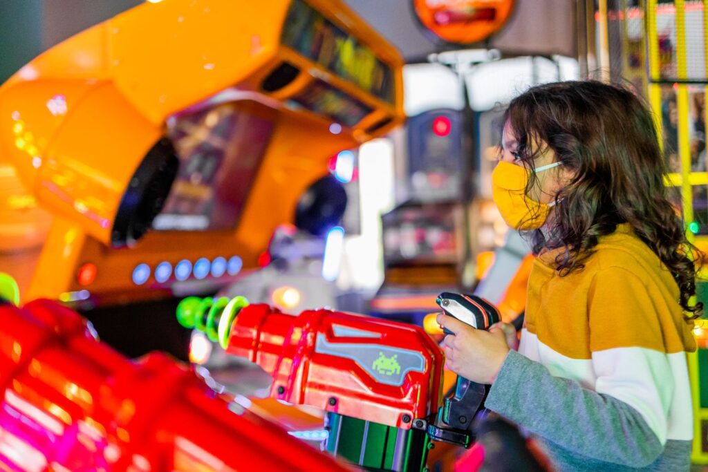 Side view of focused child in face mask playing shooting gaming machine with joysticks in arcade