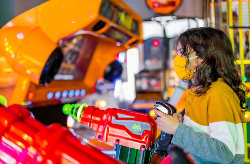 Side view of focused child in face mask playing shooting gaming machine with joysticks in arcade