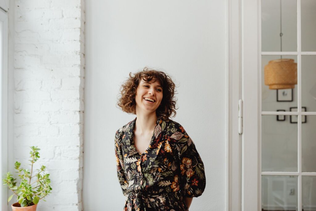 Happy woman in a floral top, standing indoors by a white wall and plant, smiling.