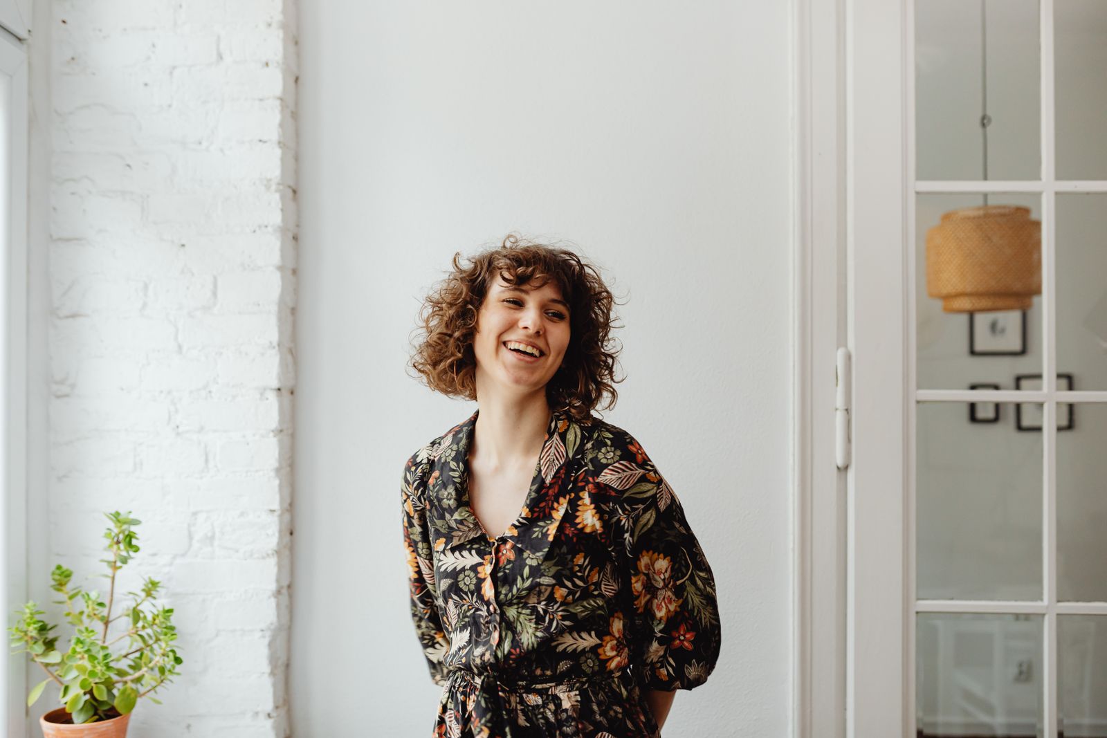 Happy woman in a floral top, standing indoors by a white wall and plant, smiling.