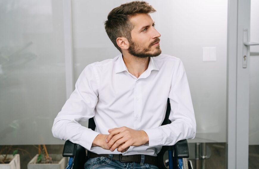 Stylish young man with a beard sitting in a wheelchair. Modern office setting, thoughtful expression.