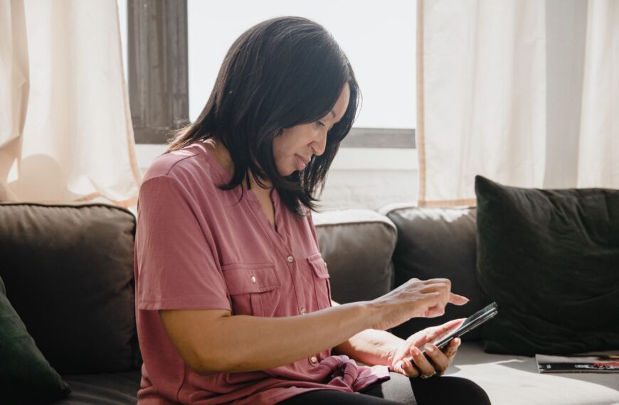 Woman in pink shirt using a smartphone while sitting on a couch, side view.