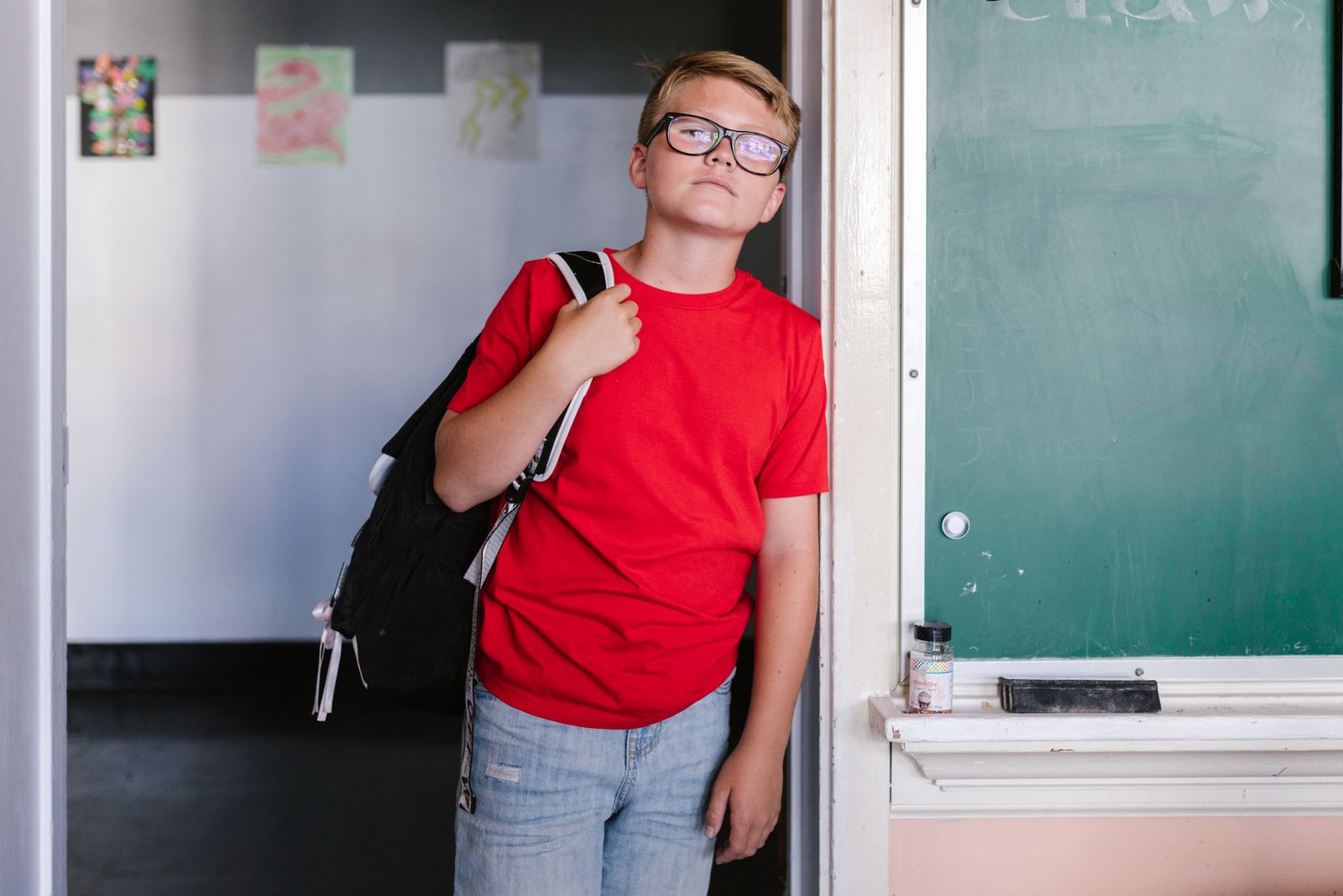 Boy in red shirt and jeans standing by a chalkboard in a classroom.