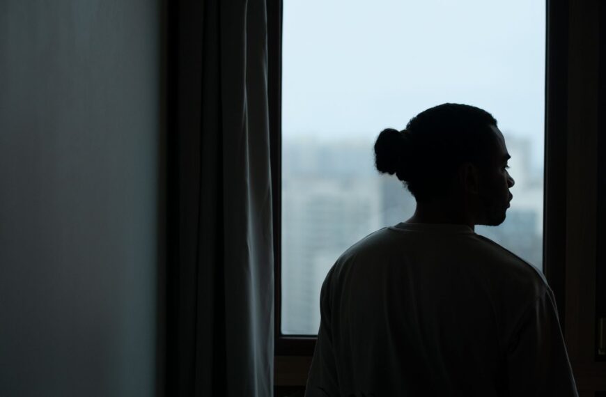 Silhouette of a man with ponytail gazing through a window with a blurred cityscape view.