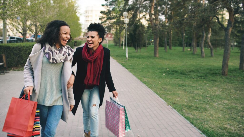 Two women happily walking with shopping bags in park.