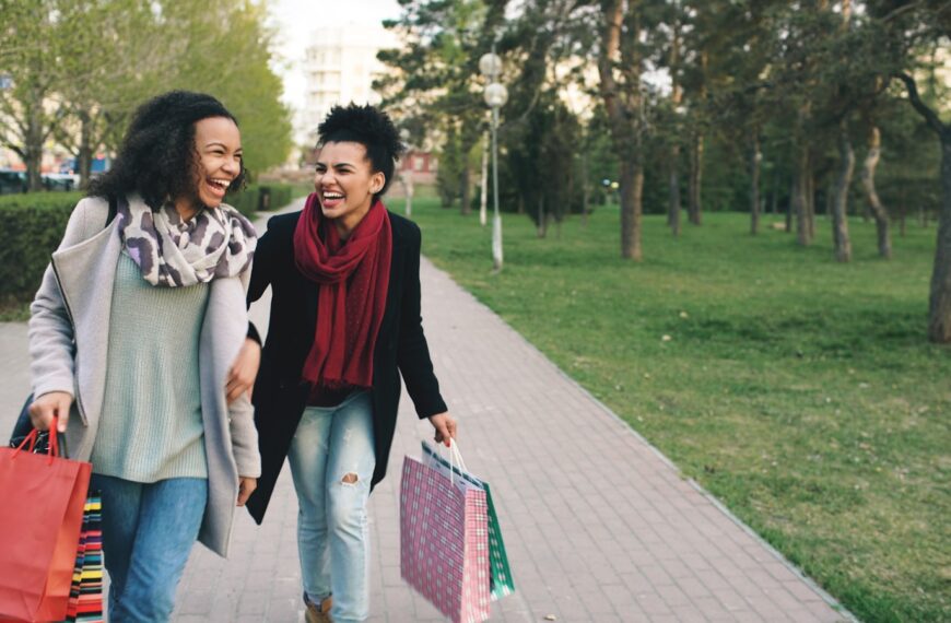Two women happily walking with shopping bags in park.