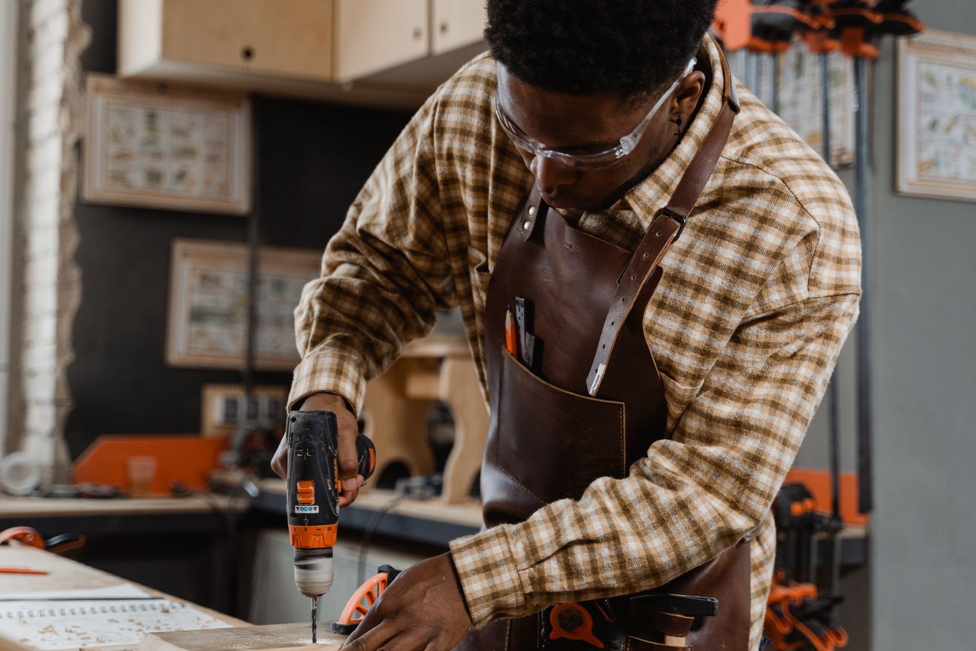Craftsman wearing safety goggles and an apron while using an electric drill on wood in a workshop