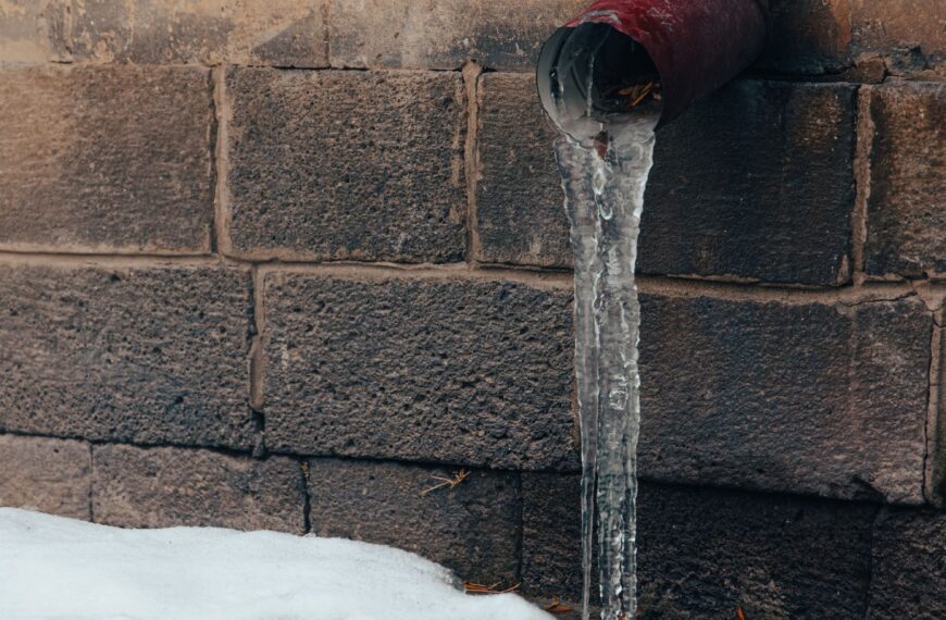 Close-up of a frozen water pipe covered in dripping icicles against an exterior stone wall in winter, illustrating the risk of burst pipes and water damage
