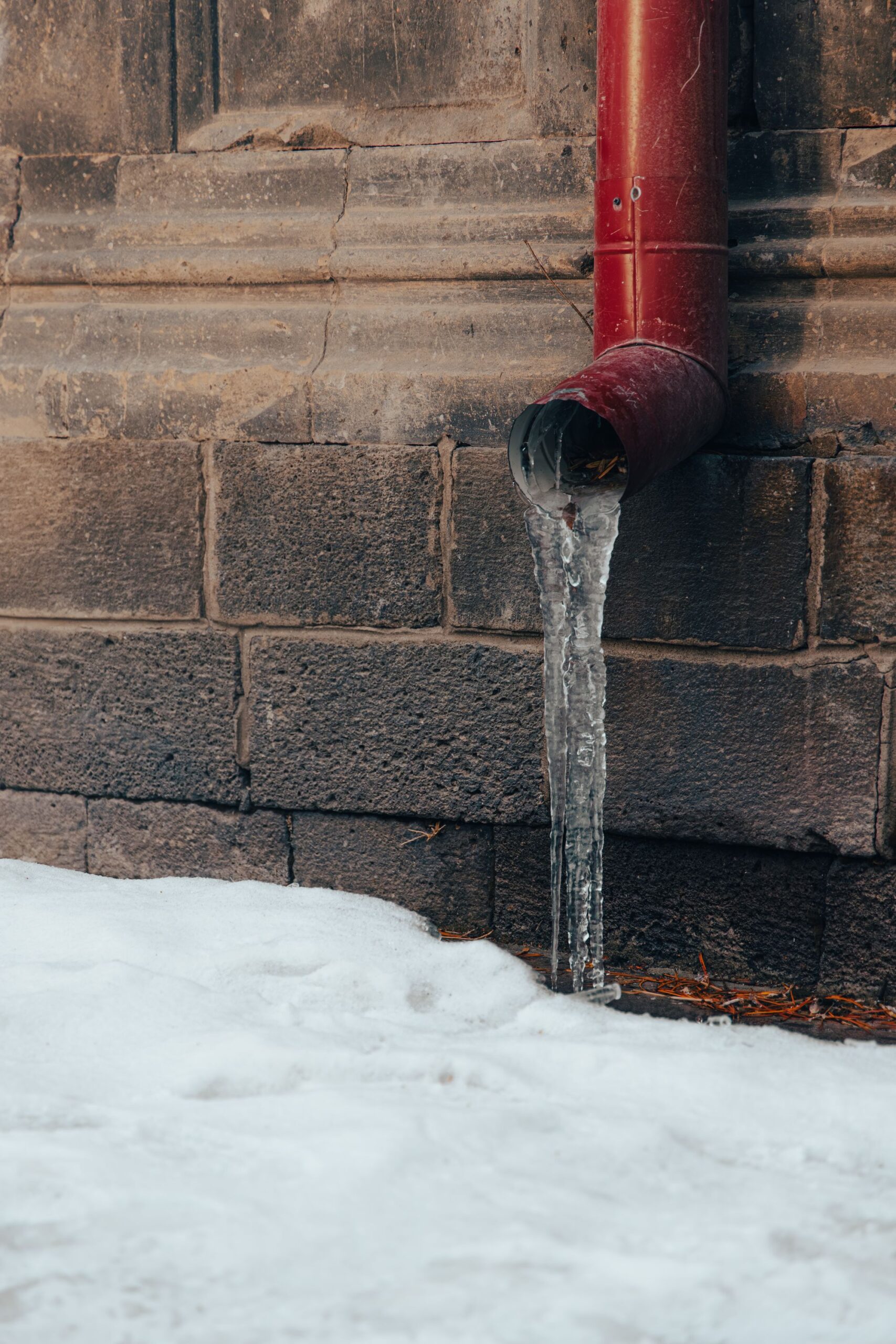 Close-up of a frozen water pipe covered in dripping icicles against an exterior stone wall in winter, illustrating the risk of burst pipes and water damage