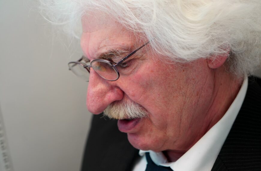 Elderly man with white hair and glasses speaking