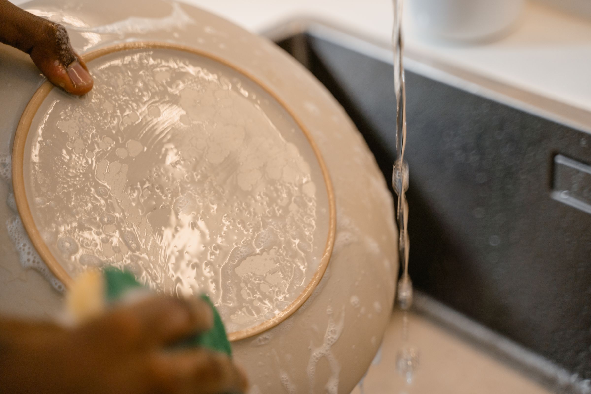 Close-up of hands hand-washing a ceramic plate with soap suds under running water at a kitchen sink