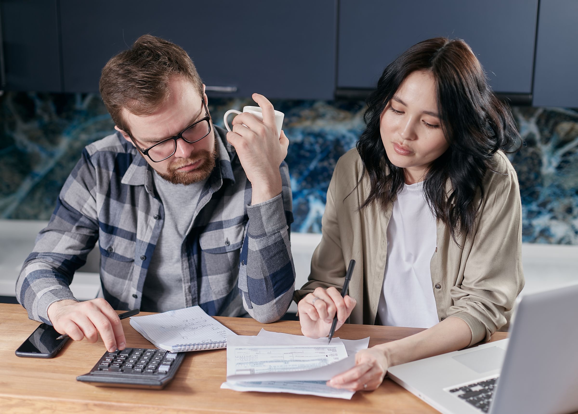Couple sitting at their kitchen table reviewing household bills and financial paperwork with a calculator and laptop