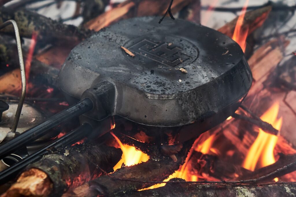 Close-up of a pie iron cooking over a campfire, surrounded by flames and logs.
