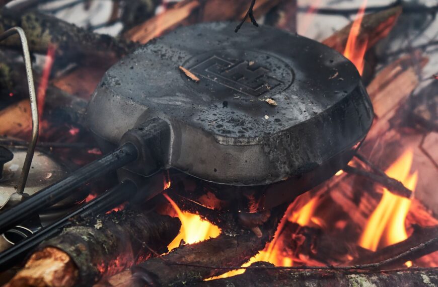 Close-up of a pie iron cooking over a campfire, surrounded by flames and logs.