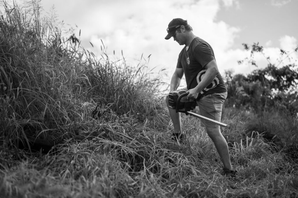 Black and white photo of a man using a chainsaw in tall grass outdoors.
