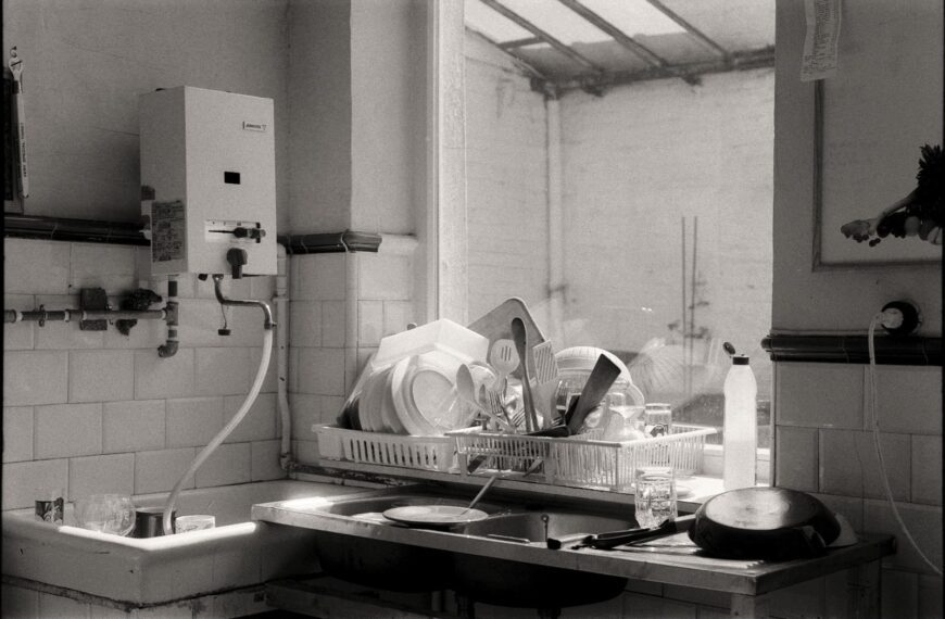Black and white image of a kitchen sink filled with dishes and cutlery by the window.