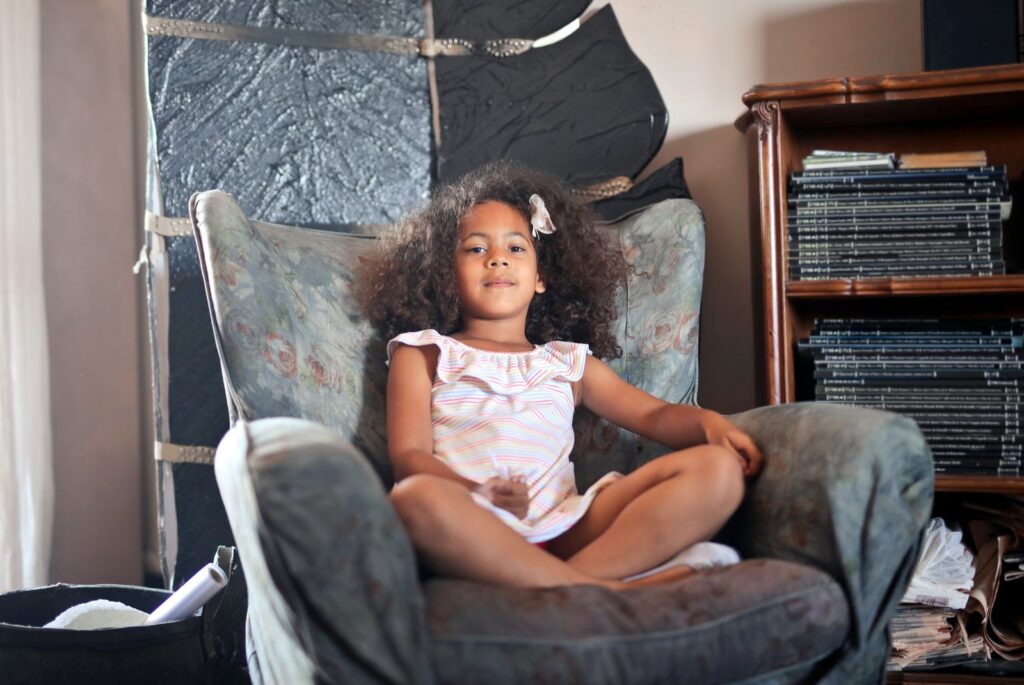 A young girl with curly hair sitting comfortably in an armchair, surrounded by books, indoors.