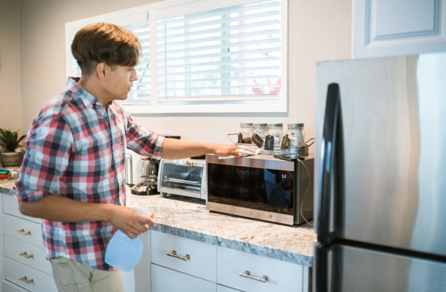 A man is cleaning a microwave in his modern kitchen using a spray bottle and cloth.