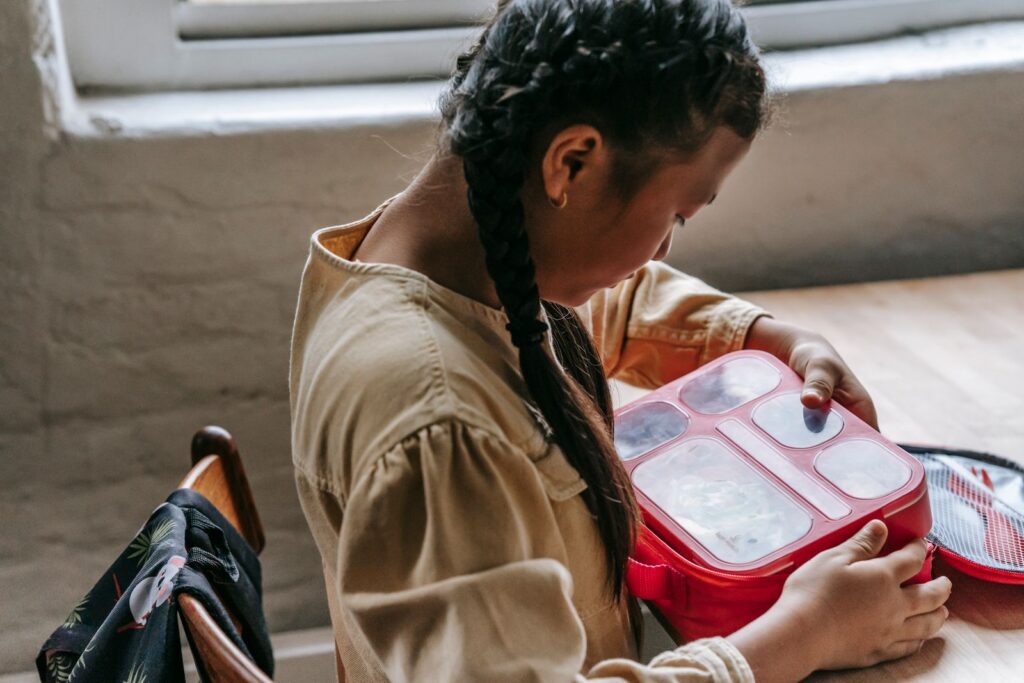 Crop teen girl in casual clothes sitting at table with plastic lunch box and preparing for breakfast