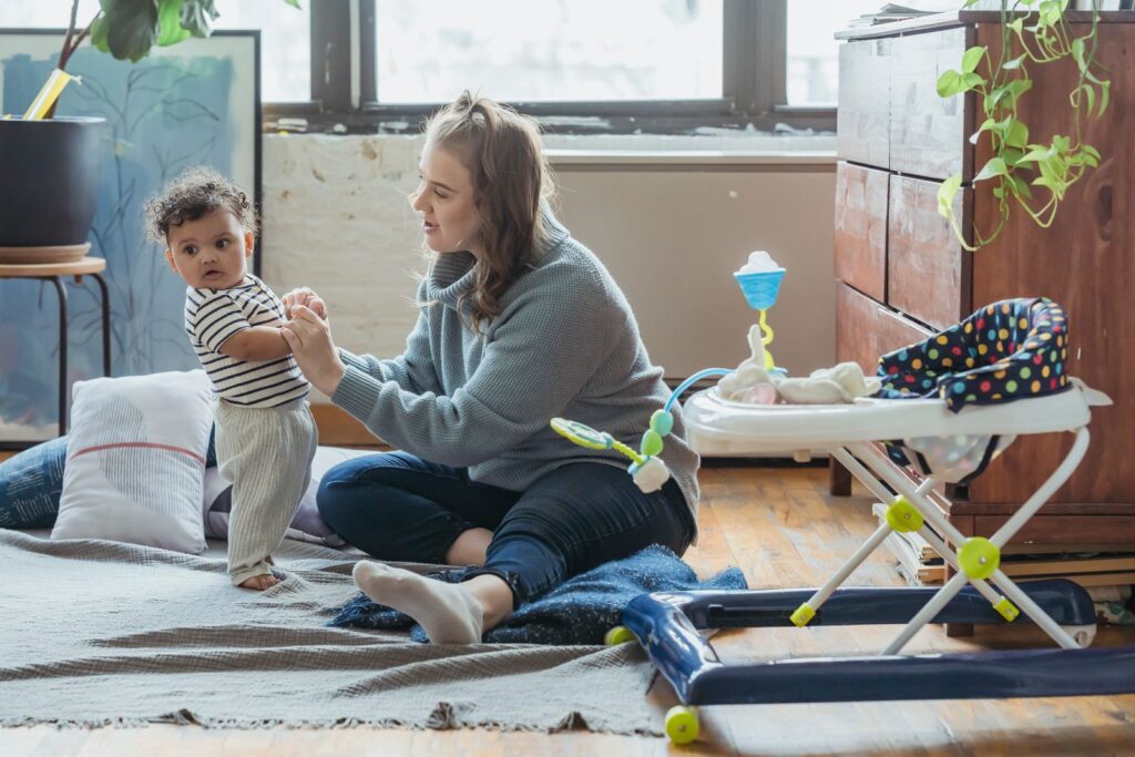 Full length positive young mother in casual clothes and adorable ethnic baby holding hands and playing together on floor in light living room