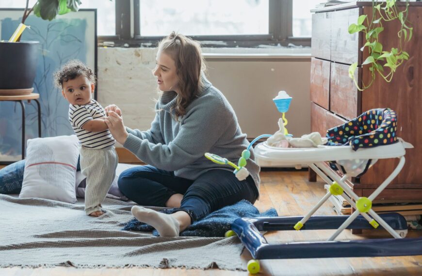 Full length positive young mother in casual clothes and adorable ethnic baby holding hands and playing together on floor in light living room