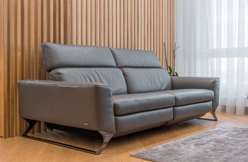 Large gray leather sofa placed in living room with carpet and windows with white curtains