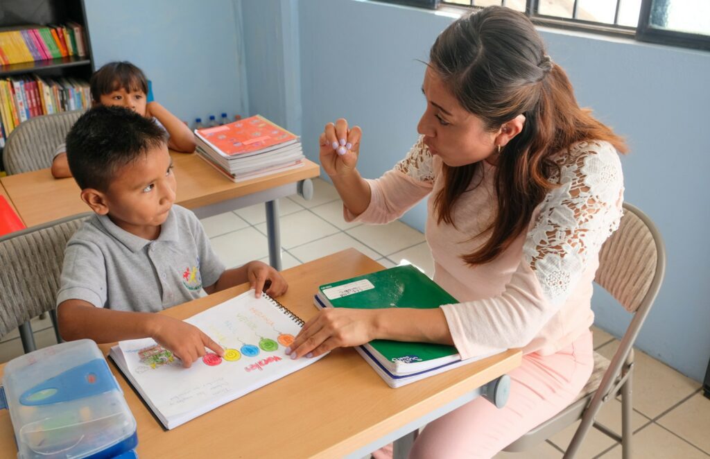 A teacher gesturing to a student pointing at a colorful workbook in a classroom