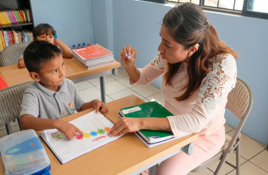 A teacher gesturing to a student pointing at a colorful workbook in a classroom
