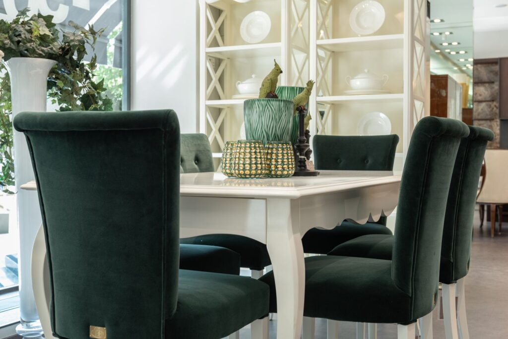 Elegant white dining table with dark green chairs near shelves with dishware in light dining room