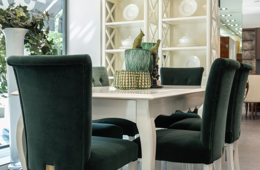 Elegant white dining table with dark green chairs near shelves with dishware in light dining room