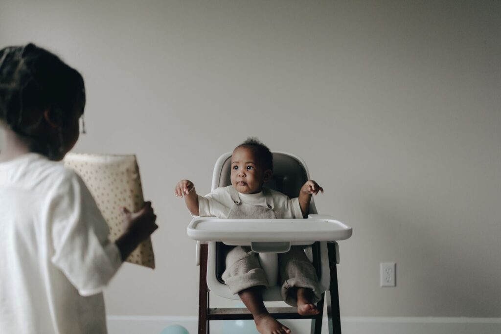 A cute baby sitting in a high chair receives a gift from a sibling indoors.