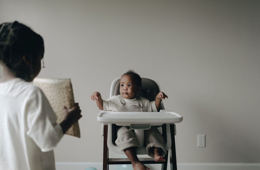 A cute baby sitting in a high chair receives a gift from a sibling indoors.