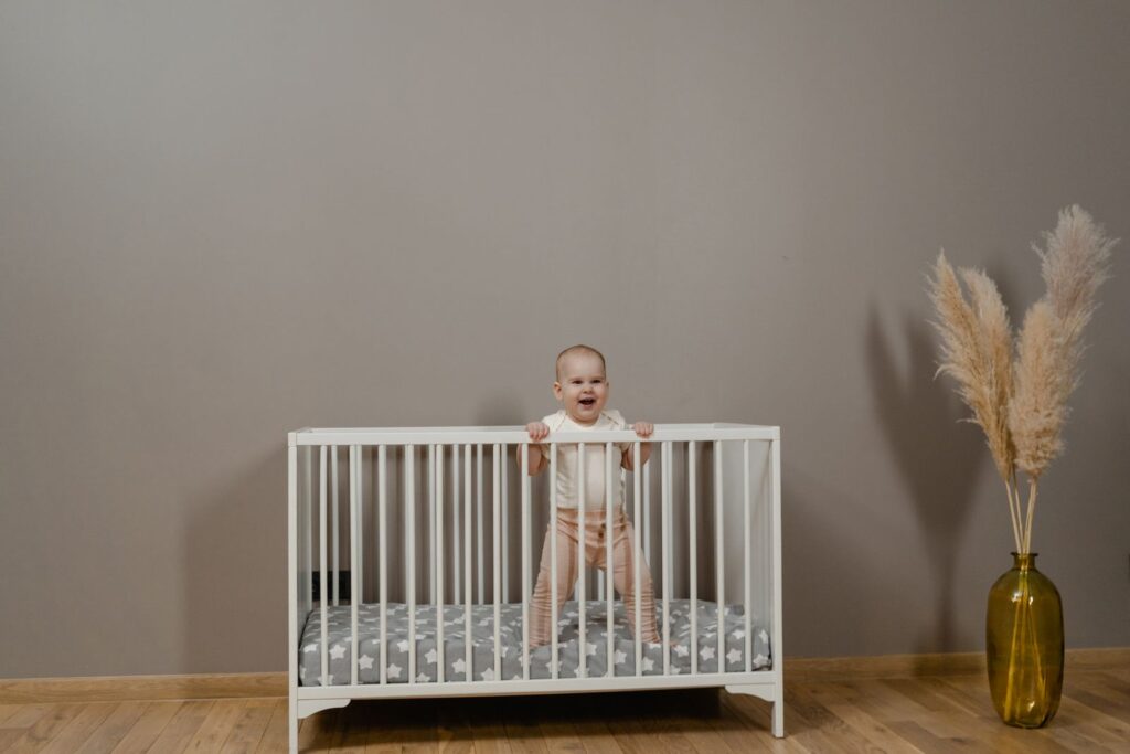 Adorable baby smiling in a white crib next to a pampas grass vase.