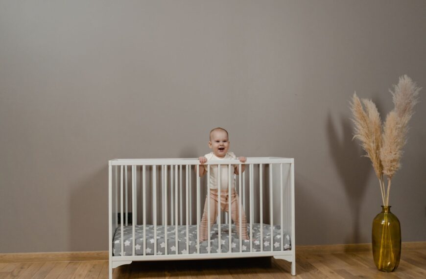 Adorable baby smiling in a white crib next to a pampas grass vase.