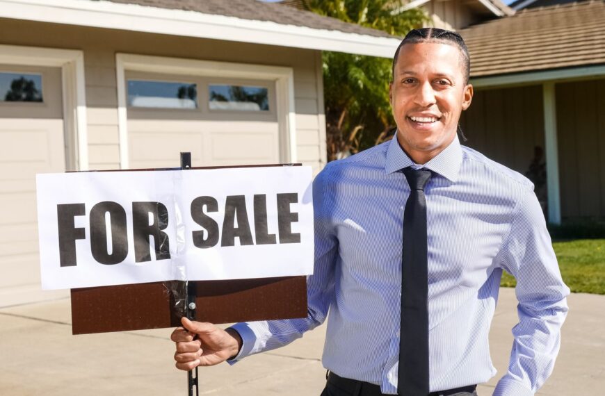 Smiling real estate agent with a for sale sign in front of a house.