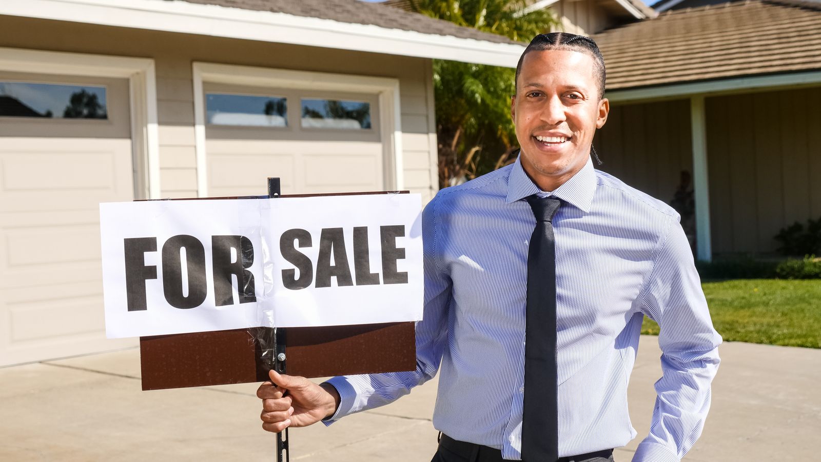 Smiling real estate agent with a for sale sign in front of a house.