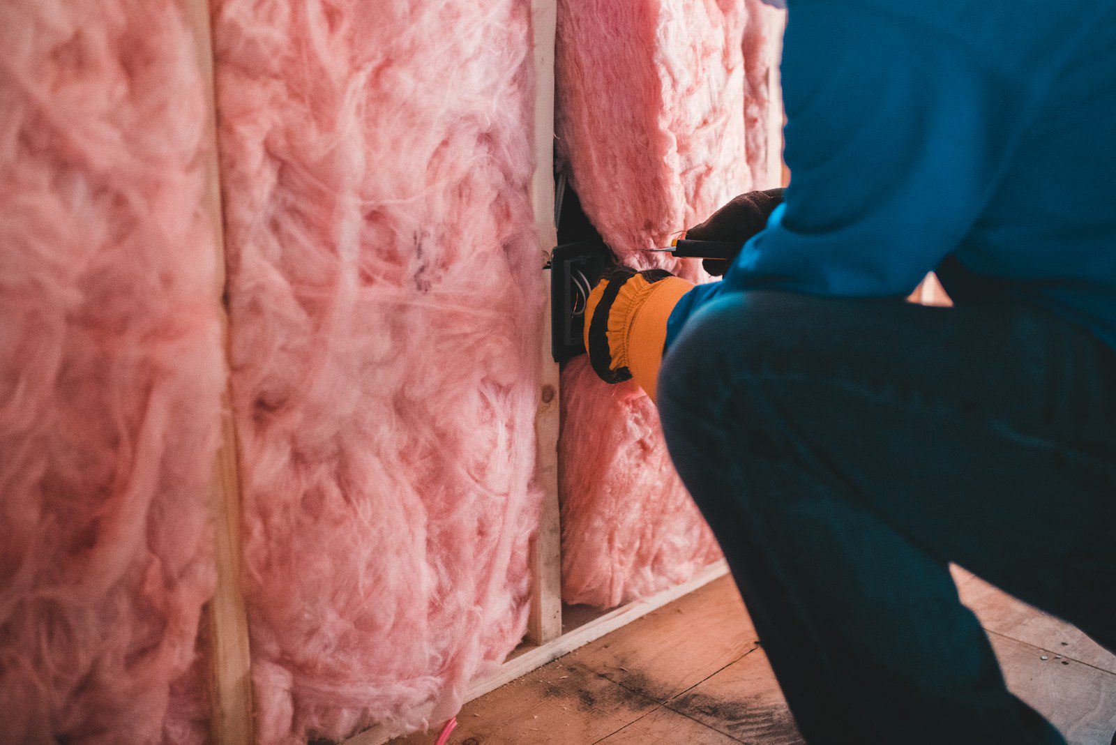 A person in a blue shirt installing electrical wiring in pink fiberglass wall insulation